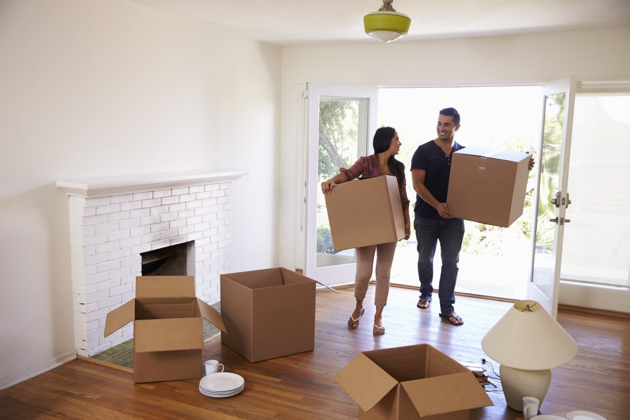 A couple carries moving boxes into a bright living room in their new home.