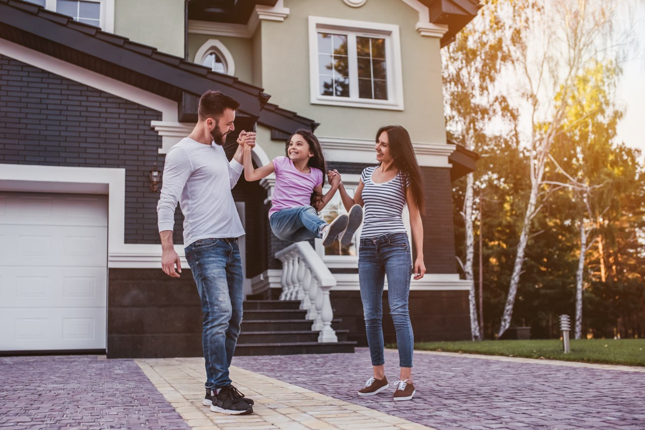 A family swings their young daughter between them in front of their modern suburban home.